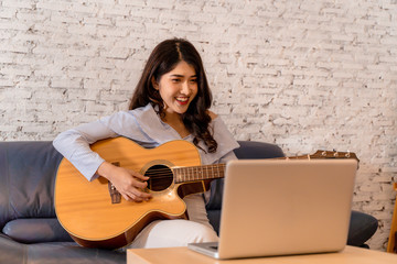 Young Asian woman practicing and learning how to play guitar on laptop computer monitor. Female...