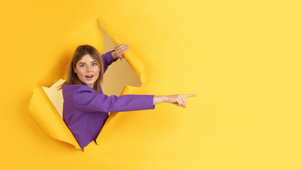 Pointing, showing. Cheerful caucasian young woman poses in torn yellow paper background, emotional and expressive. Breaking on, breakthrought. Concept of human emotions, facial expression, sales, ad. - obrazy, fototapety, plakaty