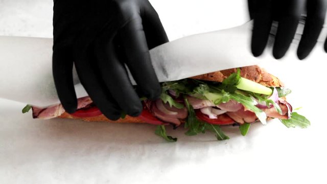 Man in black gloves wrapped in paper and big baguette sandwich meat with vegetables, time laps. Food delivery concept.