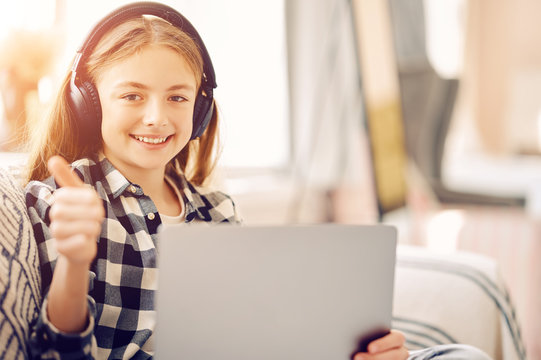 Caucasian middle schoolgirl with laptop pc and headphones at home showing thumb up and smiling. Distance education, free time, technology and internet concept