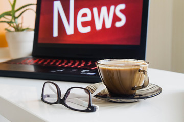 Red word news on the laptop screen. Glasses and a Cup of coffee are on the white table.