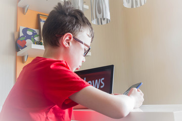 A beautiful European teen boy is viewing the news on his phone. The child is sitting at home at the table.
