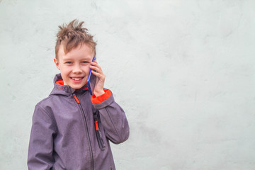 A European boy a teenager with shaggy brown hair in a blue jacket is talking on the phone and smiling. Background. Copy space.