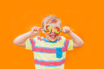A cheerful European boy covered his eyes with colored lollipops. The kid laughs. Orange joyful background.