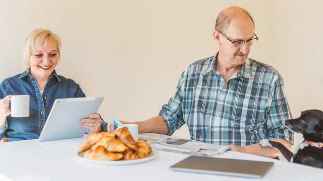 Senior couple reading news on digital tablet and old style newspaper while having breakfast in the morning - little pet (dog) asking for some food during breakfast