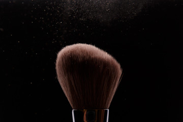 Make-up brush with beige powder explosion on black background. Creative splash. Professional powder. Cosmetics brush releasing a cloud of sparkling face powder. Copy space. Beauty and makeup concept