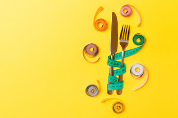 Top view composition of fork and knife with colored balled measuring tapes on yellow background with empty space for your ideas. Overweight and overeating concept