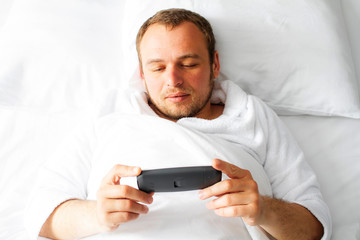 Handsome man in bed with a smartphone. Photo of a man lying in a white bed in a bathrobe.