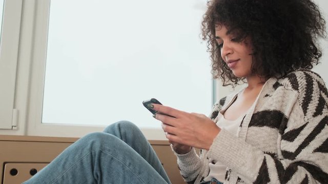 Young pleased positive african curly woman indoors at home using mobile phone