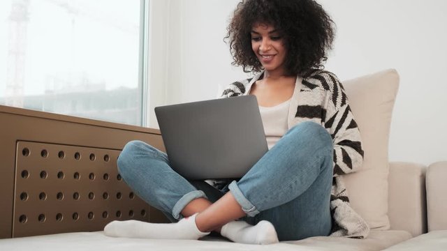 Young pleased african curly woman indoors at home using laptop computer