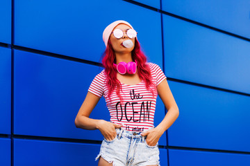 Young, bold, pretty girl, hipster with pink hair chewing gum, inflating bubbles, on a blue bright...