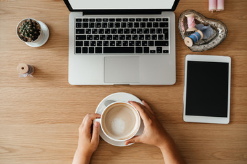 Laptop and female hands with cappuccino use the keyboard on the table in a light interior typing text and surf the internet.