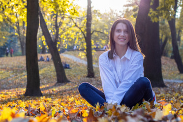 Happy girl in blue jeans and white shirt sits on fallen yellow leaves. Beautiful young woman is sitting on the ground in autumn park. Contre-jour, glares. Evening sunlight.