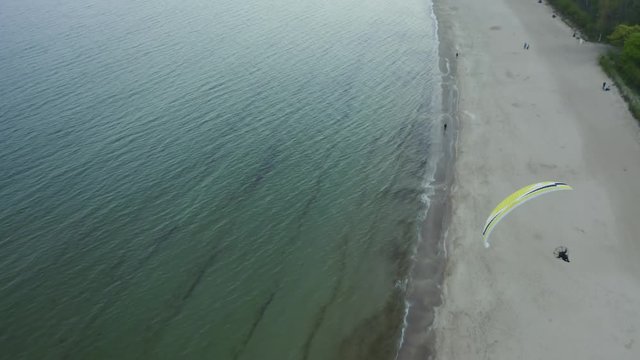 Drone flies after a paraglider flying over the sea and a sandy beach in the evening. Aerial view, motorized paraglider / paramotor above the sandy beach of the Baltic Sea