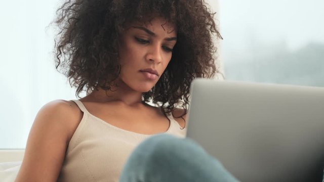 Young concentrated tired african curly woman indoors at home using laptop computer