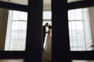 Fototapeta premium The bride and groom, European appearance. Standing at the window holding hands, looking at each other. Full height silhouette. Wedding couple.
