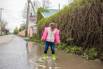 A cheerful girl is playing on the street jumping through puddles in green rubber boots. Naughty little girl jumping through puddles making big splashes