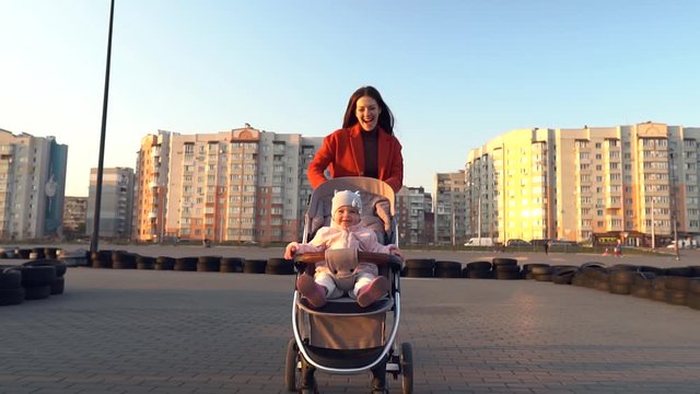 Young mom in red coat with long hair runs with a baby in stroller. Happy mother and baby. Excited toddler. Outdoor. Residential area in the background.