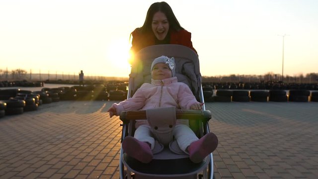 Young mother on the walk runs with a pram at the karting field. Sunset. Excited toddler, infant girl laughs. Focus goes from mom to baby. Active lifestyle. Slow motion. Sunbeams.