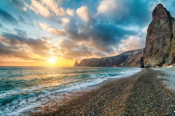 Bright sea sunset. The waves crash into the rock, lit by the warm sunset, sand and pebbles,...
