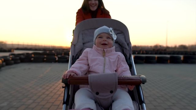 Young mother runs with a stroller at the karting field and looks at the baby. Sunset. Excited toddler, infant girl laughs. Focus goes from mom to baby. Active lifestyle. Slow motion. Sunbeams.