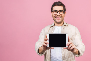 Happy young man showing blank tablet computer screen and looking at the camera isolated over pink background.