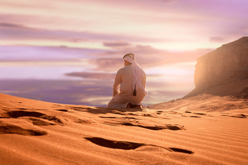 Arabic man with traditional emirates clothes sitting on kness in the UAE desert. Sunset time.