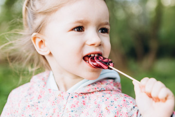 little 3-year-old girl licks a colored swirling candy on a walk in the park on a rainy summer day