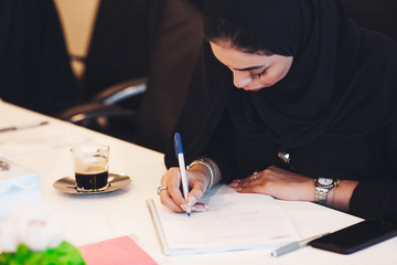 Pensive Arab woman planning working schedule writing in notebook while sitting at working place,...