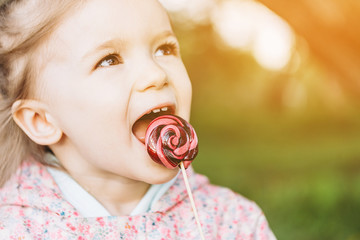 little 3-year-old girl licks a colored swirling candy on a walk in the park on a rainy summer day
