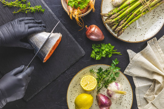 Hands in black gloves cut trout fish on black stone cutting board surrounded herbs, onion, garlic, asparagus, shrimp, prawn in ceramic plate. Black concrete table surface. Healthy seafood background