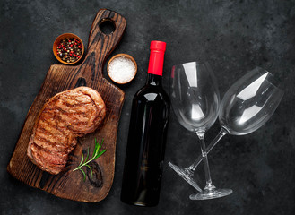 grilled beef steak with spices on a cutting board and a bottle of red wine, glasses on a stone background