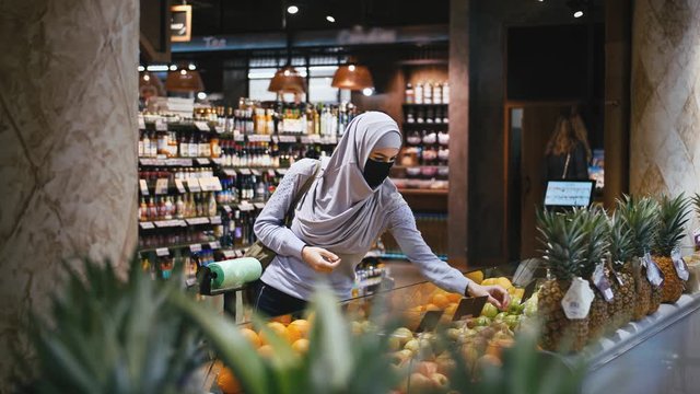 Young beautiful muslim woman in hijab and protective mask choosing fruits in the supermarket and put them in the shopping bag