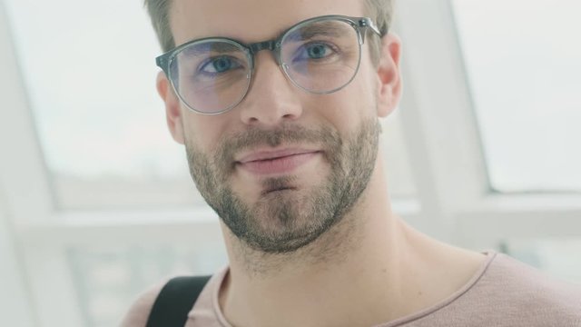 An attractive young bearded man is looking to the camera while put on his glasses while standing near the window inside