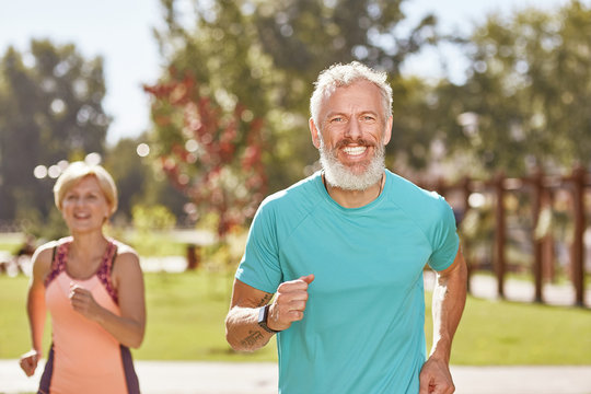 Staying Healthy And Fit. Happy Mature Man Smiling At Camera While Running Together With His Wife In The Early Morning. Selective Focus