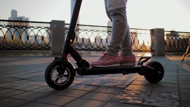 Legs of girl moves on electric scooter in city promenade during sunset