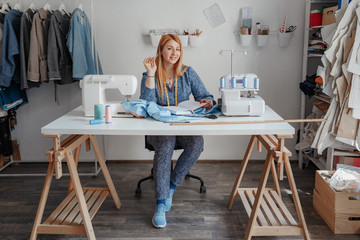 Portrait of happy young lady fashion designer smiling standing in workshop alone looking at camera. Small business owner and successful youth concept. You can see the thread, sketches, the fabric and