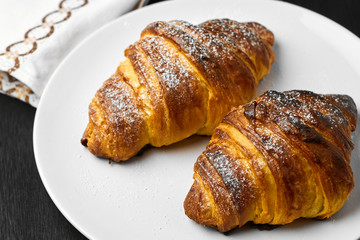 Freshly baked plain croissants on a white plate near serviette on a black wooden background. Homemade french pastry.