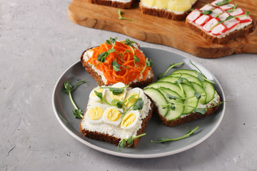Three sandwiches on toast with cream cheese, carrots, cucumber and quail eggs decorated with microgreens peas on a plate on gray concrete background, Horizontal format
