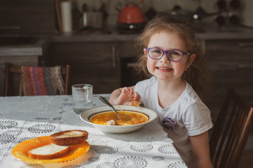 Cute little girl in glasses has breakfast in the morning at home in the kitchen. Eats toast and liquid porridge