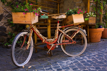 A classic red bike is decorated with wine corks and crates on a cobblestone street in Orvieto, Italy