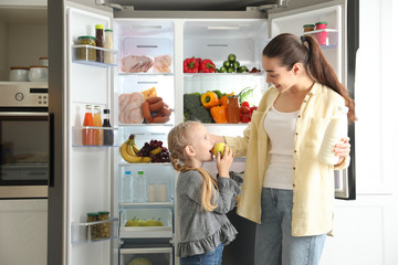 Young mother and her daughter with products near open refrigerator in kitchen