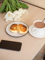 morning Breakfast with cheesecakes, phone, Cup with cacao and saucer on the table, white tulips (vertically)