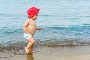 Cute adorable little blond caucasian serious pensive toddler boy in red panama walking through blue clear water waves of tropical sea or ocean beach. Vacation and travel with small children concept