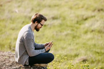 Naklejka na ściany i meble Young bearded hipster man sitting in mountains on a rock, working on the smartphone. Freelancer office in nature concept.