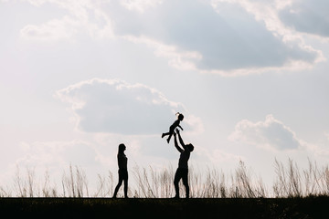 Naklejka na ściany i meble Silhouette of a family of three, on sky background. Mother, father and daughter playing together. Man throwing up little girl.