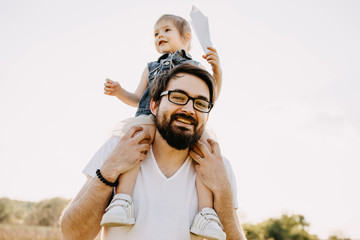 Dad playing with his daughter outdoors. Little girl sitting on father shoulders, holding a paper...