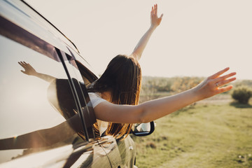 Young brunette woman sitting in a car, raising hands car outside through an open car window at sunset. Summer road trip concept.
