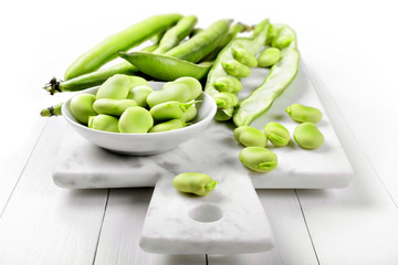 Fresh broad bean, fava beans on a marble cutting board on white wooden background, closeup.