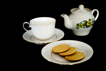 The tea set, consisting of a tea cup with a saucer, a kettle and a plate of biscuits, is located on a black background.